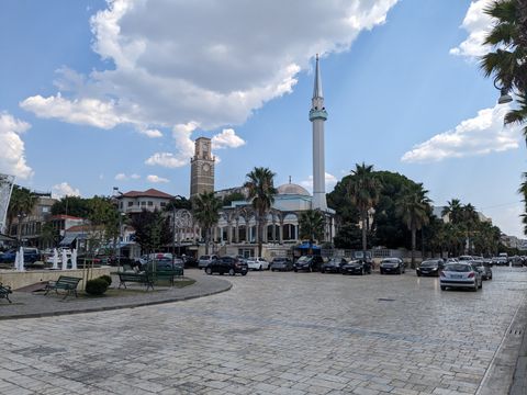 Town square of Kavaje. There's cars, palm trees, a mosque, a church tower and a fountain.